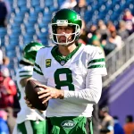 Aaron Rodgers in einer weißen Uniform der New York Jets, Nummer 8, hält einen Football, während er auf einem Feld steht. Er trägt einen grünen Helm mit Visier. Im Hintergrund sind verschwommene Zuschauer und ein blaues Stadion zu sehen. Aaron Rodgers‘ Gesichtsausdruck ist konzentriert, als er in die Kamera blickt. Diese Beschreibung wurde mit der FootballR KI automatisch generiert.