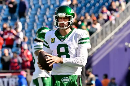 Aaron Rodgers in einer weißen Uniform der New York Jets, Nummer 8, hält einen Football, während er auf einem Feld steht. Er trägt einen grünen Helm mit Visier. Im Hintergrund sind verschwommene Zuschauer und ein blaues Stadion zu sehen. Aaron Rodgers‘ Gesichtsausdruck ist konzentriert, als er in die Kamera blickt. Diese Beschreibung wurde mit der FootballR KI automatisch generiert.