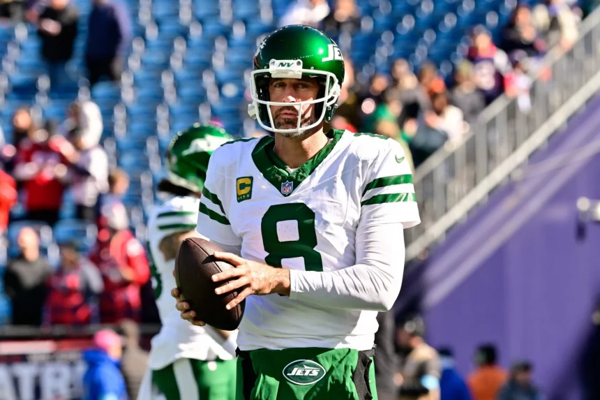Aaron Rodgers in einer weißen Uniform der New York Jets, Nummer 8, hält einen Football, während er auf einem Feld steht. Er trägt einen grünen Helm mit Visier. Im Hintergrund sind verschwommene Zuschauer und ein blaues Stadion zu sehen. Aaron Rodgers‘ Gesichtsausdruck ist konzentriert, als er in die Kamera blickt. Diese Beschreibung wurde mit der FootballR KI automatisch generiert.