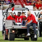 FootballR - NFL - Beim Monday Night Game wird ein Footballspieler im roten Trikot mit der Nummer 14, Chris Godwin, auf einem Wagen vom Spielfeld transportiert und signalisiert mit zwei erhobenen Fingern. Mehrere Mitarbeiter begleiten ihn, während Schiedsrichter und Zuschauer gespannt von der Tribüne aus zuschauen. Diese Beschreibung wurde automatisch generiert.