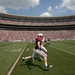 Ein American-Football-Spieler in kastanienbraunem Trikot und weißer Hose trainiert auf dem Feld und macht Dehnübungen in der Nähe der 20-Yard-Linie. Das Stadion ist voll mit Fans, viele in Rot gekleidet. Der Himmel ist teilweise bewölkt und im Hintergrund zeigt ein anderer Spieler sein Kosten-Hobby. Diese Beschreibung wurde mit der FootballR KI automatisch generiert.