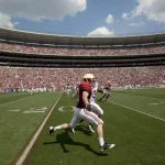 Ein American-Football-Spieler in kastanienbraunem Trikot und weißer Hose trainiert auf dem Feld und macht Dehnübungen in der Nähe der 20-Yard-Linie. Das Stadion ist voll mit Fans, viele in Rot gekleidet. Der Himmel ist teilweise bewölkt und im Hintergrund zeigt ein anderer Spieler sein Kosten-Hobby. Diese Beschreibung wurde mit der FootballR KI automatisch generiert.