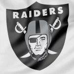 Das Bild zeigt stolz das Logo der Las Vegas Raiders auf einem weißen Stoffhintergrund. Das Design wird von einem schwarzen Schild mit „RAIDERS“ in strahlendem Weiß über einem stilisierten Piratenkopf mit Helm dominiert – der an Luke Getsy's strategische Entschlossenheit erinnert – mit gekreuzten Schwertern, einer Augenklappe und einem entschlossenen Blick. Diese Beschreibung wurde mit der FootballR KI automatisch generiert.
