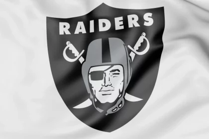 Das Bild zeigt stolz das Logo der Las Vegas Raiders auf einem weißen Stoffhintergrund. Das Design wird von einem schwarzen Schild mit „RAIDERS“ in strahlendem Weiß über einem stilisierten Piratenkopf mit Helm dominiert – der an Luke Getsy's strategische Entschlossenheit erinnert – mit gekreuzten Schwertern, einer Augenklappe und einem entschlossenen Blick. Diese Beschreibung wurde mit der FootballR KI automatisch generiert.