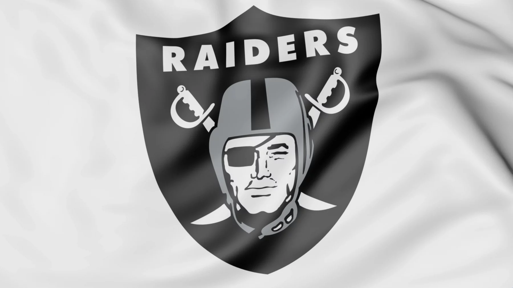 Das Bild zeigt stolz das Logo der Las Vegas Raiders auf einem weißen Stoffhintergrund. Das Design wird von einem schwarzen Schild mit „RAIDERS“ in strahlendem Weiß über einem stilisierten Piratenkopf mit Helm dominiert – der an Luke Getsy's strategische Entschlossenheit erinnert – mit gekreuzten Schwertern, einer Augenklappe und einem entschlossenen Blick. Diese Beschreibung wurde mit der FootballR KI automatisch generiert.