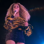 Beyoncé steht mit einem Mikrofon in der Hand und singt auf der Bühne. Sie trägt langes, lockiges Haar, ein glitzerndes, tigergestreiftes Oberteil mit Fransen und einen schwarzen Paillettenrock. Helle Bühnenlichter durchdringen die Dunkelheit und tauchen sie in eine Aura lebendiger Energie. Diese Beschreibung wurde mit der FootballR KI automatisch generiert.