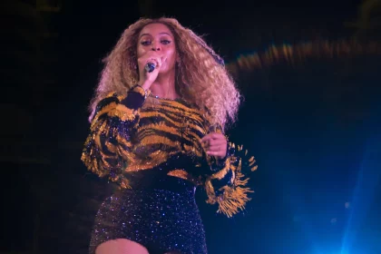 Beyoncé steht mit einem Mikrofon in der Hand und singt auf der Bühne. Sie trägt langes, lockiges Haar, ein glitzerndes, tigergestreiftes Oberteil mit Fransen und einen schwarzen Paillettenrock. Helle Bühnenlichter durchdringen die Dunkelheit und tauchen sie in eine Aura lebendiger Energie. Diese Beschreibung wurde mit der FootballR KI automatisch generiert.