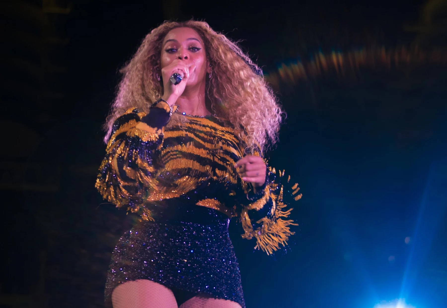 Beyoncé steht mit einem Mikrofon in der Hand und singt auf der Bühne. Sie trägt langes, lockiges Haar, ein glitzerndes, tigergestreiftes Oberteil mit Fransen und einen schwarzen Paillettenrock. Helle Bühnenlichter durchdringen die Dunkelheit und tauchen sie in eine Aura lebendiger Energie. Diese Beschreibung wurde mit der FootballR KI automatisch generiert.