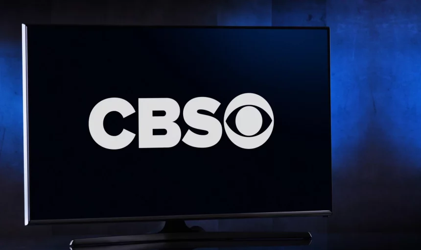 Ein Breitbildfernseher zeigt einen dunkelblauen Bildschirm mit dem weißen CBS-Logo, während die Spannung vor dem bevorstehenden Spiel Chiefs vs. Bills steigt. Die fettgedruckten „CBS“-Buchstaben und das ikonische Augensymbol fallen auf, während der schwach beleuchtete Hintergrund das elegante Design des Fernsehers betont. Diese Beschreibung wurde mit der FootballR KI automatisch generiert.