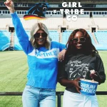 Zwei Frauen posieren aufgeregt in einem Stadion. Eine trägt eine Sonnenbrille, einen blauen Panthers-Pullover und Jeans und hebt die Hand. Die andere trägt eine Sonnenbrille, einen grauen Carolina Panthers-Pullover aus der NFL-Kollektion und einen leichten Rock und hält Popcorn in der Hand. Im oberen Hintergrund sind ein Logo und der Text „Girl Tribe Co.“ zu sehen. Diese Beschreibung wurde mit der FootballR KI automatisch generiert.