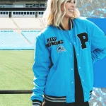 Eine Frau steht lächelnd in einem Stadion und trägt stolz eine hellblaue Collegejacke aus der NFL-Kollektion der Carolina Panthers mit den Aufnähern „P“ und „Keep Pounding“. Ihr schulterlanges blondes Haar passt perfekt zu ihrem schwarzen Outfit. Hinter ihr blicken leere Sitze auf das leuchtend grüne Feld. Diese Beschreibung wurde mit der FootballR KI automatisch generiert.