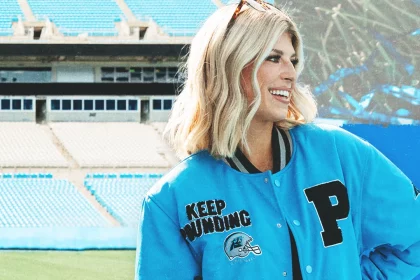Eine Frau steht lächelnd in einem Stadion und trägt stolz eine hellblaue Collegejacke aus der NFL-Kollektion der Carolina Panthers mit den Aufnähern „P“ und „Keep Pounding“. Ihr schulterlanges blondes Haar passt perfekt zu ihrem schwarzen Outfit. Hinter ihr blicken leere Sitze auf das leuchtend grüne Feld. Diese Beschreibung wurde mit der FootballR KI automatisch generiert.