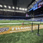 Thanksgiving 2026: Die NFL Thanksgiving Games erwachen auf einem farbenfrohen Footballfeld in einem großen Stadion zum Leben, auf dessen Rasen „THANKSGIVING“ prangt. Helle Lichter erhellen die Szene, während sich die Menschen an den Seitenlinien versammeln und große Bildschirme Touchdowns und Spielberichte übertragen. Eine schwarze Absperrung aus Seilen markiert den vorderen Bereich. Diese Beschreibung wurde mit der FootballR KI automatisch generiert.