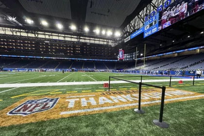 Thanksgiving 2026: Die NFL Thanksgiving Games erwachen auf einem farbenfrohen Footballfeld in einem großen Stadion zum Leben, auf dessen Rasen „THANKSGIVING“ prangt. Helle Lichter erhellen die Szene, während sich die Menschen an den Seitenlinien versammeln und große Bildschirme Touchdowns und Spielberichte übertragen. Eine schwarze Absperrung aus Seilen markiert den vorderen Bereich. Diese Beschreibung wurde mit der FootballR KI automatisch generiert.