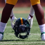 Ein Footballspieler mit grünen Socken und weißen Stollen hockt auf einem Feld und hält einen Helm mit einem gelben Streifen und dem Etikett „SPARTANS“. Die helle, sonnige Umgebung hebt die gelben Shorts des Spielers hervor und betont seine Bereitschaft und Konzentration auf dem Feld. Diese Beschreibung wurde mit der FootballR KI automatisch generiert.