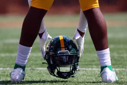 Ein Footballspieler mit grünen Socken und weißen Stollen hockt auf einem Feld und hält einen Helm mit einem gelben Streifen und dem Etikett „SPARTANS“. Die helle, sonnige Umgebung hebt die gelben Shorts des Spielers hervor und betont seine Bereitschaft und Konzentration auf dem Feld. Diese Beschreibung wurde mit der FootballR KI automatisch generiert.