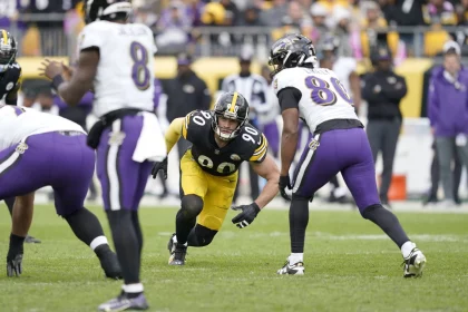 Ein Footballspieler in schwarz-gelber Uniform mit der Nummer 90 ist bereit, das Spielfeld zu betreten. Er ist von Spielern in weiß-lila Uniformen umgeben, einer trägt die Nummer 8. Diese actiongeladene Szene spielt sich inmitten einer lebhaften Menge während der NFL Woche 11 ab und ist eine weitere spannende Geschichte der Saison. Diese Beschreibung wurde mit der FootballR KI automatisch generiert.
