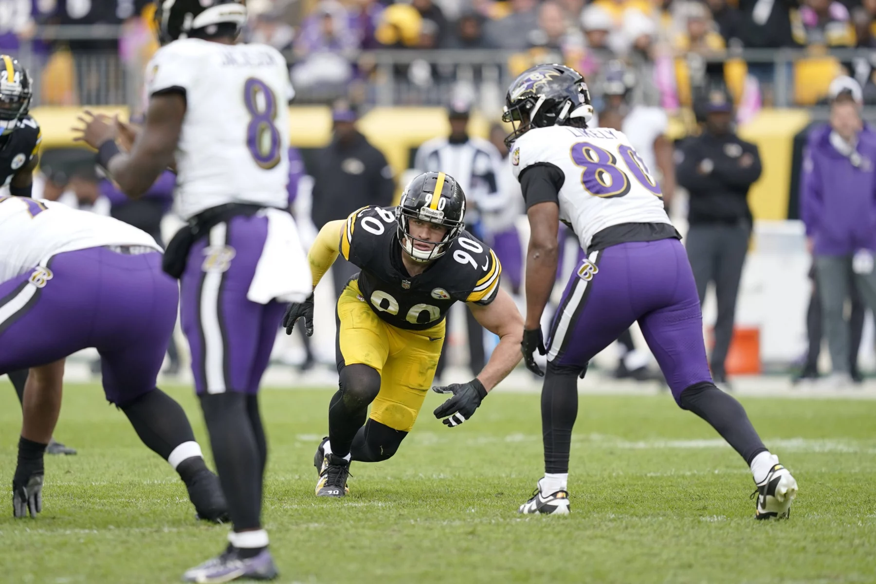 Ein Footballspieler in schwarz-gelber Uniform mit der Nummer 90 ist bereit, das Spielfeld zu betreten. Er ist von Spielern in weiß-lila Uniformen umgeben, einer trägt die Nummer 8. Diese actiongeladene Szene spielt sich inmitten einer lebhaften Menge während der NFL Woche 11 ab und ist eine weitere spannende Geschichte der Saison. Diese Beschreibung wurde mit der FootballR KI automatisch generiert.
