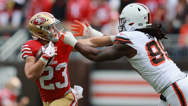Christian McCaffrey in einer rot-goldenen 49ers-Uniform hält den Ball, während er von Za'Darius Smith in einer weißen Browns-Uniform angegriffen wird. Der 49ers-Spieler versucht, Smith während eines Spiels mit seinem Arm abzuwehren, während im Hintergrund rot gekleidete Zuschauer zu sehen sind. Diese Beschreibung wurde mit der FootballR KI automatisch generiert.