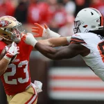 Christian McCaffrey in einer rot-goldenen 49ers-Uniform hält den Ball, während er von Za'Darius Smith in einer weißen Browns-Uniform angegriffen wird. Der 49ers-Spieler versucht, Smith während eines Spiels mit seinem Arm abzuwehren, während im Hintergrund rot gekleidete Zuschauer zu sehen sind. Diese Beschreibung wurde mit der FootballR KI automatisch generiert.