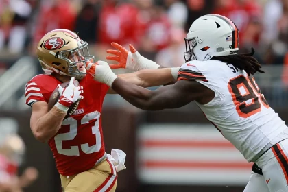 Christian McCaffrey in einer rot-goldenen 49ers-Uniform hält den Ball, während er von Za'Darius Smith in einer weißen Browns-Uniform angegriffen wird. Der 49ers-Spieler versucht, Smith während eines Spiels mit seinem Arm abzuwehren, während im Hintergrund rot gekleidete Zuschauer zu sehen sind. Diese Beschreibung wurde mit der FootballR KI automatisch generiert.