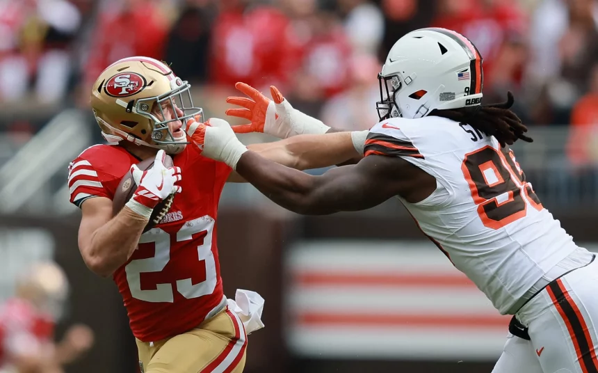 Christian McCaffrey in einer rot-goldenen 49ers-Uniform hält den Ball, während er von Za'Darius Smith in einer weißen Browns-Uniform angegriffen wird. Der 49ers-Spieler versucht, Smith während eines Spiels mit seinem Arm abzuwehren, während im Hintergrund rot gekleidete Zuschauer zu sehen sind. Diese Beschreibung wurde mit der FootballR KI automatisch generiert.