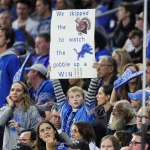 Ein junger Fan bei einem Footballspiel hält ein Schild hoch, auf dem steht: „Wir haben auf den Truthahn verzichtet, um den Detroit Lions dabei zuzusehen, wie sie einen SIEG einfahren!!!“ Die Menge um das Kind herum ist auf das Spiel konzentriert, viele tragen blaue Teamkleidung. Die Atmosphäre ist lebhaft und voller Zuschauer. Diese Beschreibung wurde mit der FootballR KI automatisch generiert.