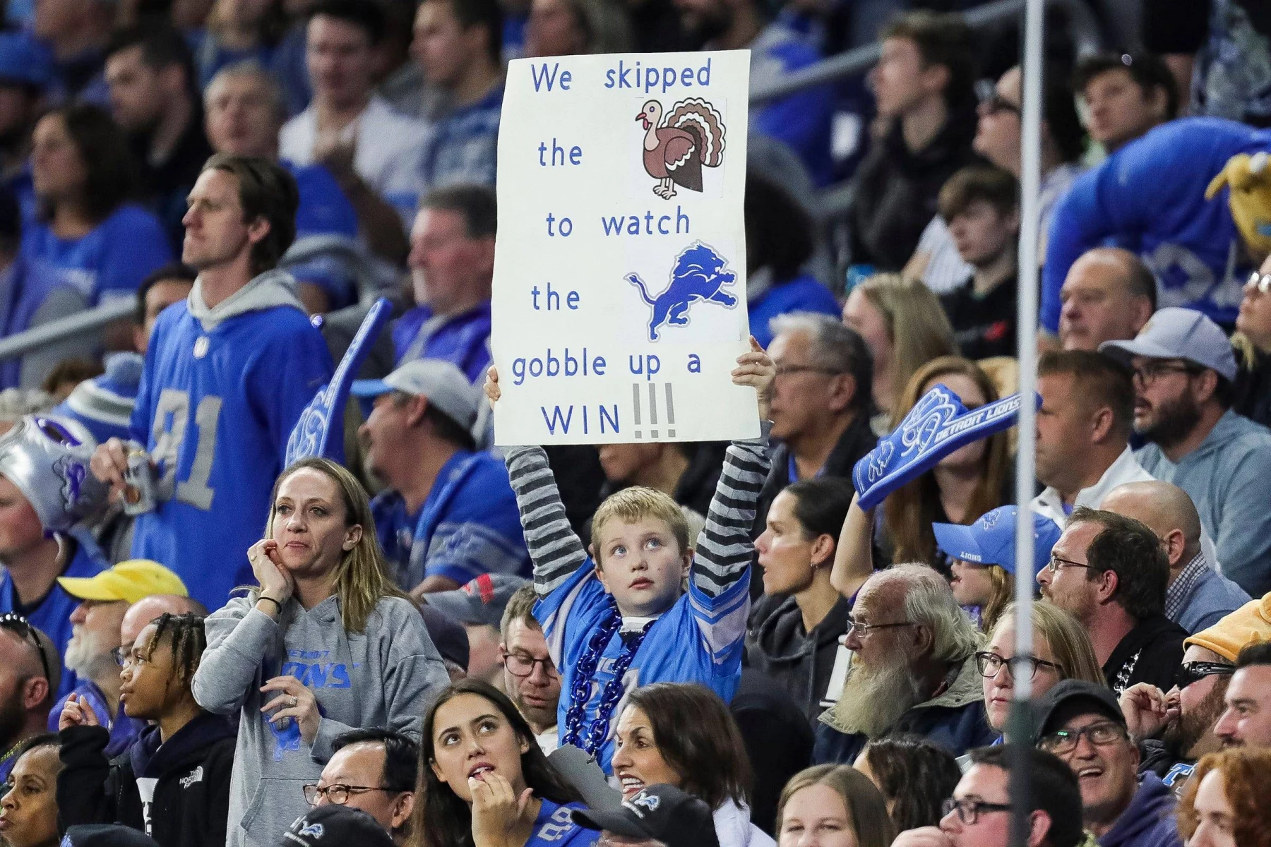 Ein junger Fan bei einem Footballspiel hält ein Schild hoch, auf dem steht: „Wir haben auf den Truthahn verzichtet, um den Detroit Lions dabei zuzusehen, wie sie einen SIEG einfahren!!!“ Die Menge um das Kind herum ist auf das Spiel konzentriert, viele tragen blaue Teamkleidung. Die Atmosphäre ist lebhaft und voller Zuschauer. Diese Beschreibung wurde mit der FootballR KI automatisch generiert.