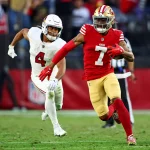 Charvarius Ward, in roter Uniform und goldenem Helm, rennt mit dem Ball über das Rasenfeld, dicht gefolgt von einem entschlossenen Spieler in weißem Trikot und Helm. Im Hintergrund sind Zuschauer und eine offizielle Tribüne verschwommen. Diese Beschreibung wurde mit der FootballR KI automatisch generiert.