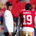 Ein Footballtrainer in einem weißen Langarmshirt und einer schwarzen Kappe, Kyle Shanahan, hält einen Football und lächelt Deebo Samuel an. Der Spieler, der ein rotes 49ers-Trikot mit der Nummer 19 und „Samuel“ trägt, steht dem Trainer auf dem Feld gegenüber, während im Hintergrund eine verschwommene Menge roter Fans jubelt. Diese Beschreibung wurde mit der FootballR KI automatisch generiert.
