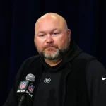 Joe Douglas, mit Bart, erscheint in einem schwarzen Kapuzenpullover und einer ärmellosen Jacke am Podium. Er spricht selbstbewusst zum Publikum in ein Mikrofon mit NFL-Logo, das vor einem dunklen Vorhang im Hintergrund steht. Diese Beschreibung wurde mit der FootballR KI automatisch generiert.