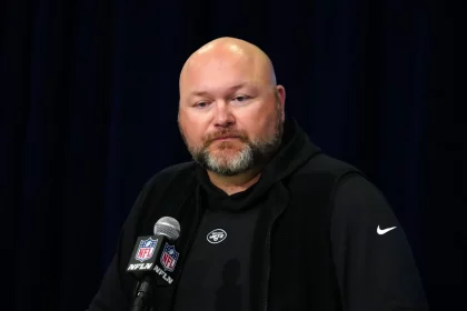 Joe Douglas, mit Bart, erscheint in einem schwarzen Kapuzenpullover und einer ärmellosen Jacke am Podium. Er spricht selbstbewusst zum Publikum in ein Mikrofon mit NFL-Logo, das vor einem dunklen Vorhang im Hintergrund steht. Diese Beschreibung wurde mit der FootballR KI automatisch generiert.