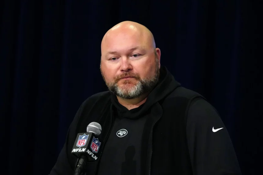 Joe Douglas, mit Bart, erscheint in einem schwarzen Kapuzenpullover und einer ärmellosen Jacke am Podium. Er spricht selbstbewusst zum Publikum in ein Mikrofon mit NFL-Logo, das vor einem dunklen Vorhang im Hintergrund steht. Diese Beschreibung wurde mit der FootballR KI automatisch generiert.