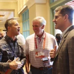 Drei Männer unterhalten sich auf einer Konferenz. John Harbaugh links trägt ein kariertes Hemd und hält ein Telefon und ein Notizbuch in der Hand. Der Mann in der Mitte in einem gestreiften Poloshirt blickt Jim Harbaugh rechts an, der einen Anzug trägt. Konferenzabzeichen hängen an ihren Hälsen wie Chargers-Fans, die ihr Team repräsentieren. Über ihnen funkeln Kronleuchter. Diese Beschreibung wurde mit der FootballR KI automatisch generiert.