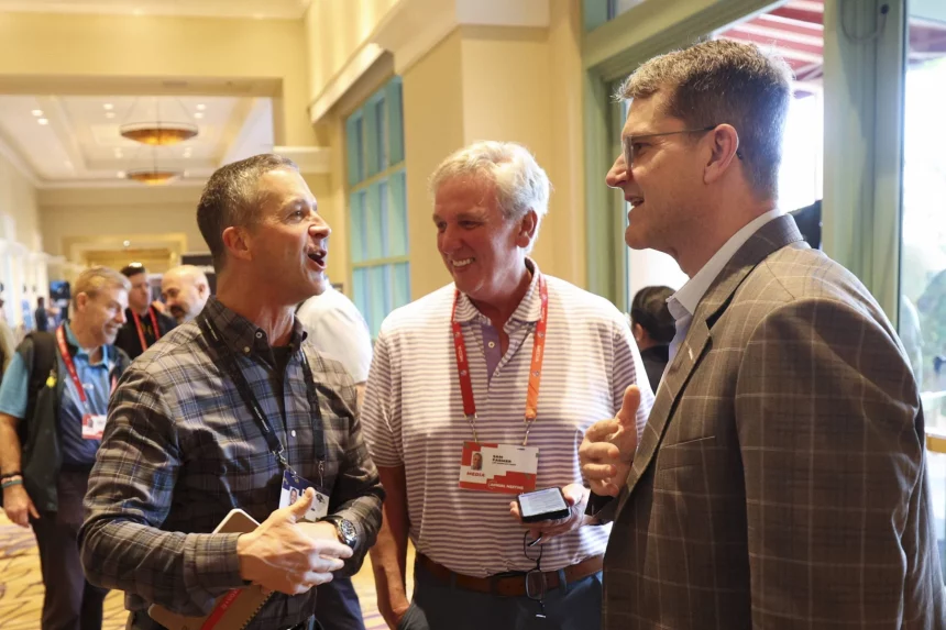 Drei Männer unterhalten sich auf einer Konferenz. John Harbaugh links trägt ein kariertes Hemd und hält ein Telefon und ein Notizbuch in der Hand. Der Mann in der Mitte in einem gestreiften Poloshirt blickt Jim Harbaugh rechts an, der einen Anzug trägt. Konferenzabzeichen hängen an ihren Hälsen wie Chargers-Fans, die ihr Team repräsentieren. Über ihnen funkeln Kronleuchter. Diese Beschreibung wurde mit der FootballR KI automatisch generiert.