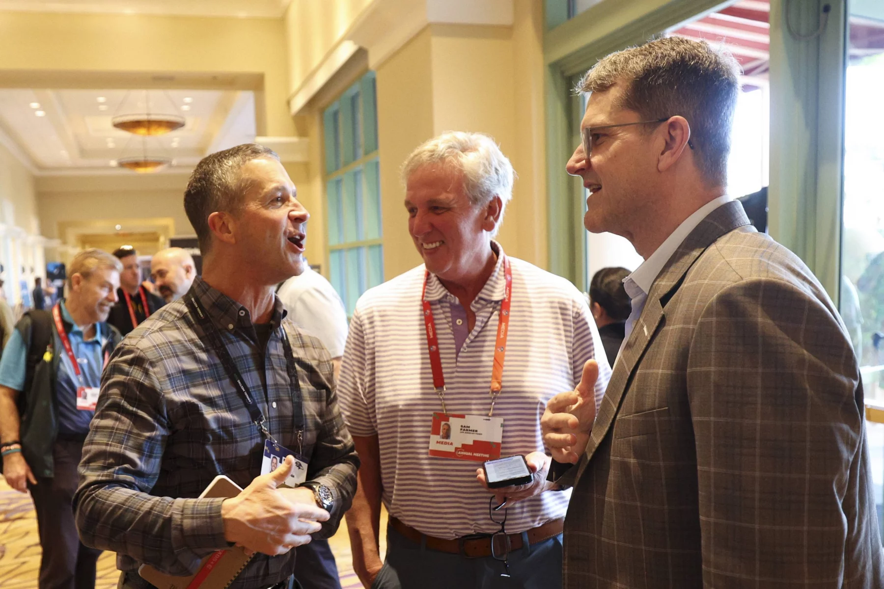 Drei Männer unterhalten sich auf einer Konferenz. John Harbaugh links trägt ein kariertes Hemd und hält ein Telefon und ein Notizbuch in der Hand. Der Mann in der Mitte in einem gestreiften Poloshirt blickt Jim Harbaugh rechts an, der einen Anzug trägt. Konferenzabzeichen hängen an ihren Hälsen wie Chargers-Fans, die ihr Team repräsentieren. Über ihnen funkeln Kronleuchter. Diese Beschreibung wurde mit der FootballR KI automatisch generiert.