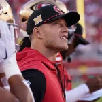Ein Footballspieler in einer roten San Francisco 49ers-Weste und einer schwarzen Kappe steht während eines Spiels an der Seitenlinie. Er blickt aufmerksam auf das Spielfeld, wie Christian McCaffrey, der Spielzüge analysiert. Zwei Teamkollegen mit Helmen stehen in der Nähe, und im Hintergrund schreibt ein Trainer auf ein Klemmbrett. Das Stadion ist voller Zuschauer. Diese Beschreibung wurde mit der FootballR KI automatisch generiert.