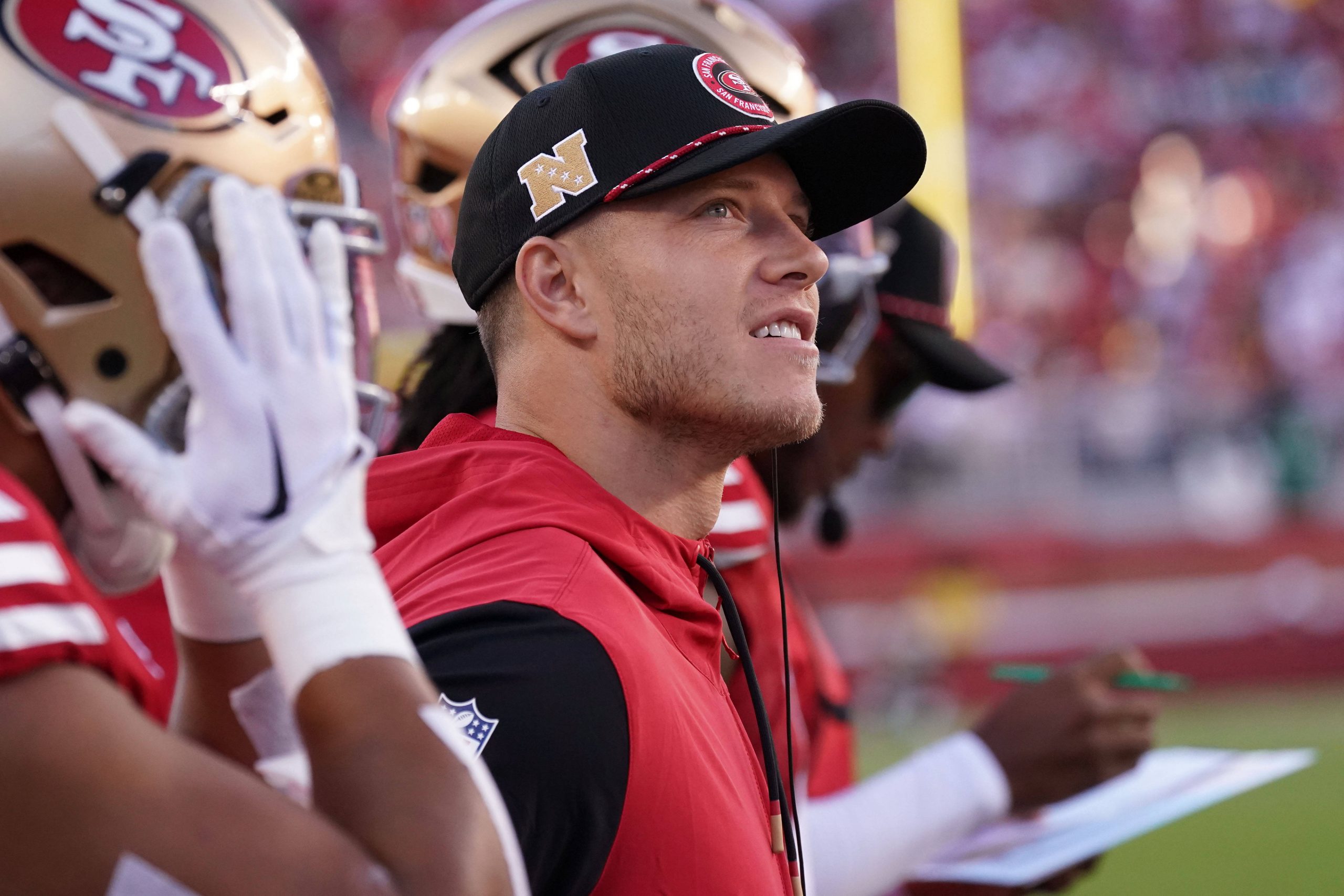 Ein Footballspieler in einer roten San Francisco 49ers-Weste und einer schwarzen Kappe steht während eines Spiels an der Seitenlinie. Er blickt aufmerksam auf das Spielfeld, wie Christian McCaffrey, der Spielzüge analysiert. Zwei Teamkollegen mit Helmen stehen in der Nähe, und im Hintergrund schreibt ein Trainer auf ein Klemmbrett. Das Stadion ist voller Zuschauer. Diese Beschreibung wurde mit der FootballR KI automatisch generiert.