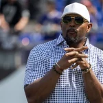 Ray Lewis mit weißer Baseballkappe, Sonnenbrille und einem blau-weiß karierten Hemd steht im Freien. Mit Spitzbart und gefalteten Händen posiert er vor einem leicht verschwommenen Hintergrund aus Menschen und Stadionsitzen. Diese Beschreibung wurde mit der FootballR KI automatisch generiert.