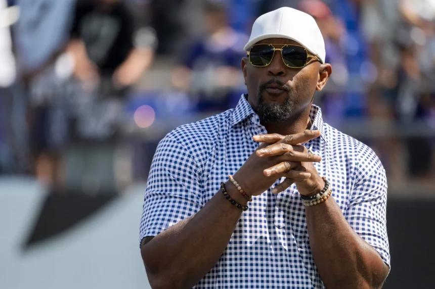 Ray Lewis mit weißer Baseballkappe, Sonnenbrille und einem blau-weiß karierten Hemd steht im Freien. Mit Spitzbart und gefalteten Händen posiert er vor einem leicht verschwommenen Hintergrund aus Menschen und Stadionsitzen. Diese Beschreibung wurde mit der FootballR KI automatisch generiert.