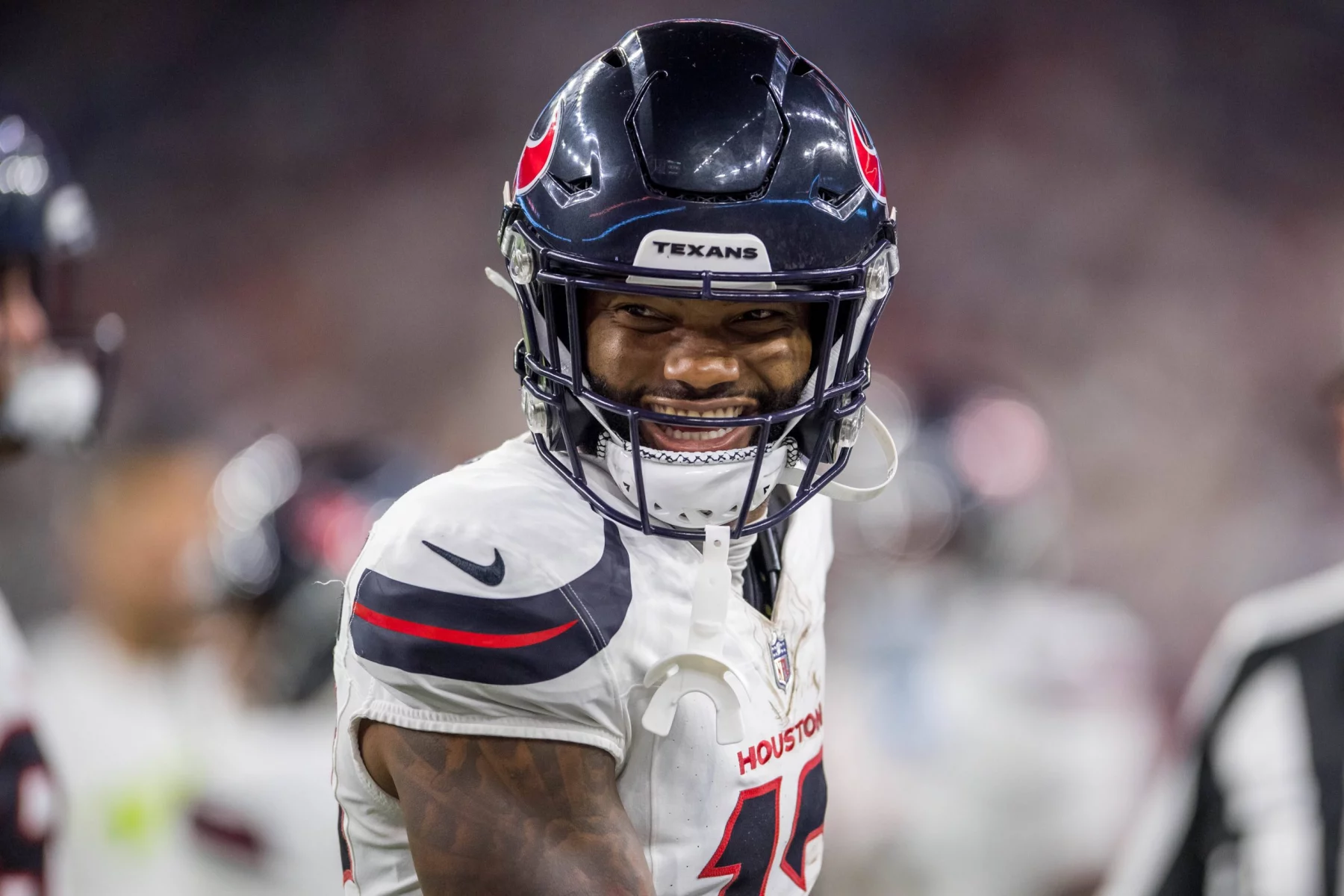 Nico Collins, der Footballspieler mit dem dunklen Helm und der Aufschrift „Texans“, lächelt auf dem Feld. Er trägt eine weiße Uniform mit der Aufschrift „Houston“ auf der Vorderseite, hervorgehoben durch schwarze und rote Akzente. Im Hintergrund sind verschwommene Figuren und Stadionlichter zu sehen. Diese Beschreibung wurde mit der FootballR KI automatisch generiert.