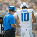 Ein Footballspieler in weißer Uniform mit der „10“ hört aufmerksam zu, während Trainer Jim Harbaugh, der Kopfhörer trägt und in Blau gekleidet ist, Anweisungen gibt. Ein Offizieller und eine verschwommene Menge runden die lebhafte Spieltagsatmosphäre ab. Diese Beschreibung wurde mit der FootballR KI automatisch generiert.