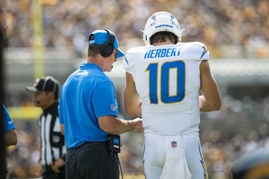 Ein Footballspieler in weißer Uniform mit der „10“ hört aufmerksam zu, während Trainer Jim Harbaugh, der Kopfhörer trägt und in Blau gekleidet ist, Anweisungen gibt. Ein Offizieller und eine verschwommene Menge runden die lebhafte Spieltagsatmosphäre ab. Diese Beschreibung wurde mit der FootballR KI automatisch generiert.