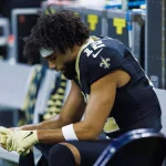 Auf der Bank sitzt Chris Olave in schwarz-goldener Uniform. Er lehnt sich nach vorne und stützt seine Ellbogen auf die Knie. Er trägt das Trikot mit der Nummer 12 und ein Stirnband. Seine behandschuhten Hände sind gefaltet. Im Hintergrund sind Sportgeräte zu sehen. Diese Beschreibung wurde mit der FootballR KI automatisch generiert.