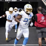 J.K. Dobbins in weiß-blauen Uniformen verlassen einen Tunnel auf das Spielfeld. An der Spitze der Gruppe steht die Nummer 27, dicht gefolgt von der Nummer 0. Ein Mann in einer roten Weste mit der Aufschrift „NFL Television“ auf dem Rücken steht an der Seite und hält eine Kamera, während über ihm auf einem Banner „ESTD: 1869“ steht. Diese Beschreibung wurde mit der FootballR KI automatisch generiert.