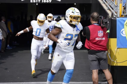 J.K. Dobbins in weiß-blauen Uniformen verlassen einen Tunnel auf das Spielfeld. An der Spitze der Gruppe steht die Nummer 27, dicht gefolgt von der Nummer 0. Ein Mann in einer roten Weste mit der Aufschrift „NFL Television“ auf dem Rücken steht an der Seite und hält eine Kamera, während über ihm auf einem Banner „ESTD: 1869“ steht. Diese Beschreibung wurde mit der FootballR KI automatisch generiert.
