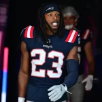 Kyle Dugger in einer dunkelblauen Uniform der New England Patriots mit der Nummer 23, möglicherweise Dugger, schlendert durch einen Tunnel. Seine langen Haare sind von einem Stirnband zurückgehalten und er trägt einen blauen Armstulpen. Rote und blaue Lichter erhellen die Szene, und hinter ihm ist ein weiterer Spieler leicht zu erkennen. Diese Beschreibung wurde mit der FootballR KI automatisch generiert.