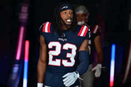 Kyle Dugger in einer dunkelblauen Uniform der New England Patriots mit der Nummer 23, möglicherweise Dugger, schlendert durch einen Tunnel. Seine langen Haare sind von einem Stirnband zurückgehalten und er trägt einen blauen Armstulpen. Rote und blaue Lichter erhellen die Szene, und hinter ihm ist ein weiterer Spieler leicht zu erkennen. Diese Beschreibung wurde mit der FootballR KI automatisch generiert.