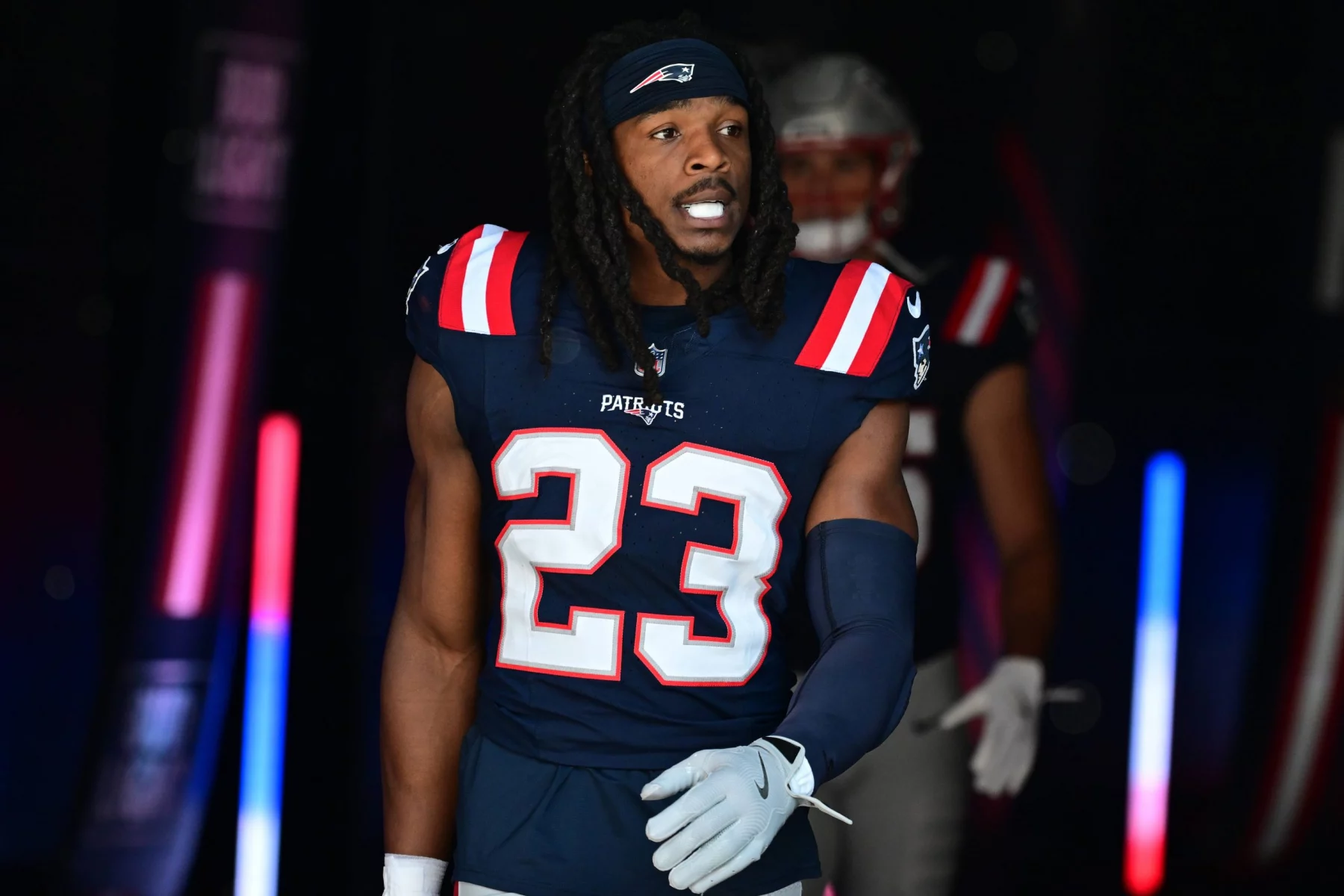 Kyle Dugger in einer dunkelblauen Uniform der New England Patriots mit der Nummer 23, möglicherweise Dugger, schlendert durch einen Tunnel. Seine langen Haare sind von einem Stirnband zurückgehalten und er trägt einen blauen Armstulpen. Rote und blaue Lichter erhellen die Szene, und hinter ihm ist ein weiterer Spieler leicht zu erkennen. Diese Beschreibung wurde mit der FootballR KI automatisch generiert.