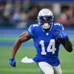 Ein Footballspieler, DK Metcalf, in einem blauen Trikot mit der Nummer 14 und einem silbernen Helm, sprintet über das Feld und hält den Ball mit seinem rechten Arm. Der unscharfe Hintergrund zeigt ein Publikum und Stadionlichter, was darauf schließen lässt, dass ein spannendes Seahawks-Spiel im Gange ist. Diese Beschreibung wurde mit der FootballR KI automatisch generiert.